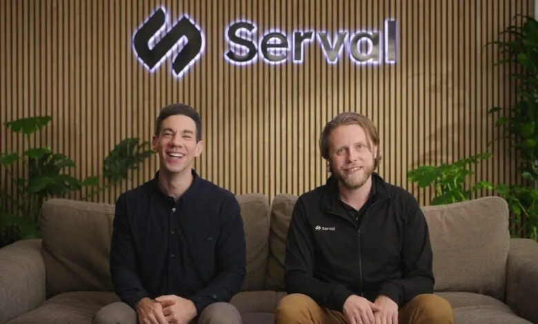 Two men smiling on a couch in front of the Serval logo and wood-paneled wall.