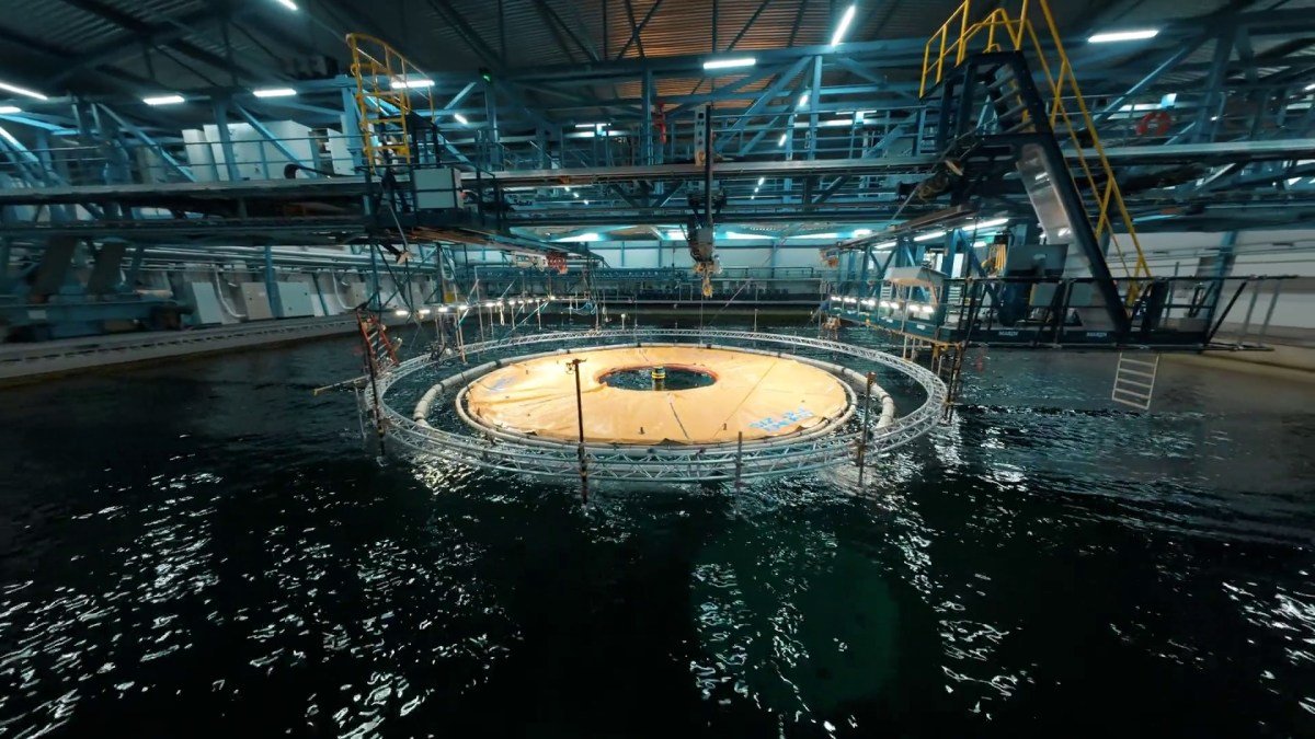 Circular floating platform prototype in a large indoor water basin with industrial lighting.