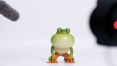 A small, green toy frog with large eyes and orange feet sits on a white surface, facing the camera.