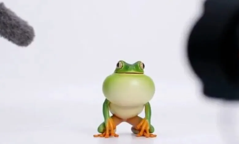 A small, green toy frog with large eyes and orange feet sits on a white surface, facing the camera.