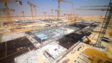 Aerial view of a massive construction site with numerous cranes and steel structures under a clear blue sky.
