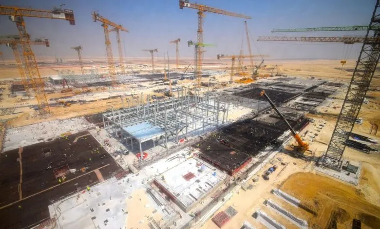 Aerial view of a massive construction site with numerous cranes and steel structures under a clear blue sky.
