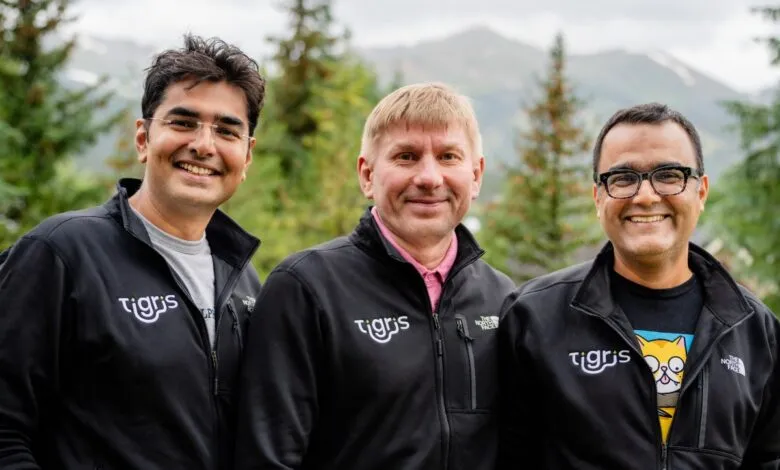 Three smiling men in black jackets with the Tigris logo, standing outdoors with trees and mountains in the background.