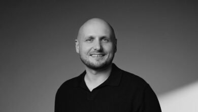 Bald man with a beard and a black collared shirt smiles at the camera.