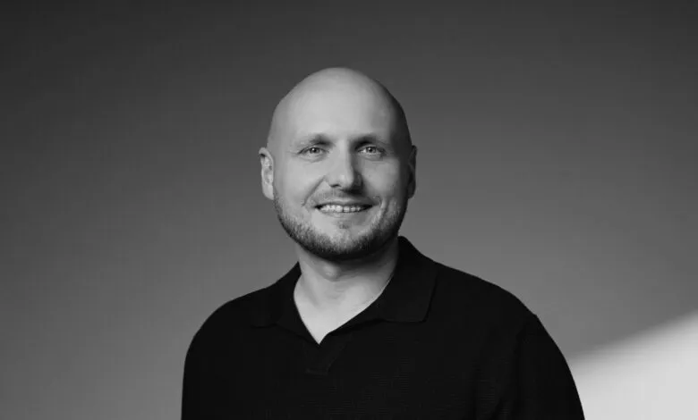 Bald man with a beard and a black collared shirt smiles at the camera.