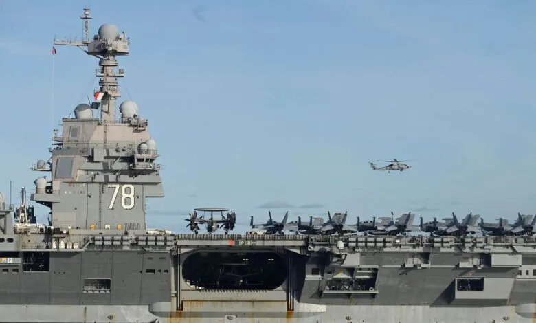 USS Gerald R. Ford aircraft carrier with fighter jets and a helicopter in flight.