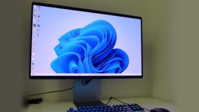 A modern computer monitor displaying the Windows 11 logo and a keyboard with blue backlighting.
