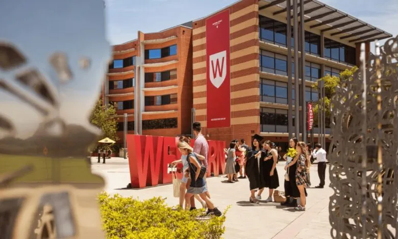 Students in graduation attire posing near large red 'WESTERN' letters at university.