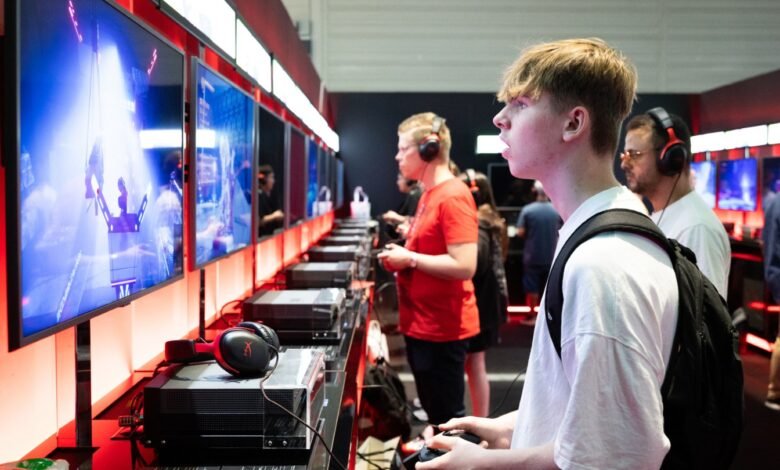 Young man playing video games with headphones on, surrounded by other gamers and screens.