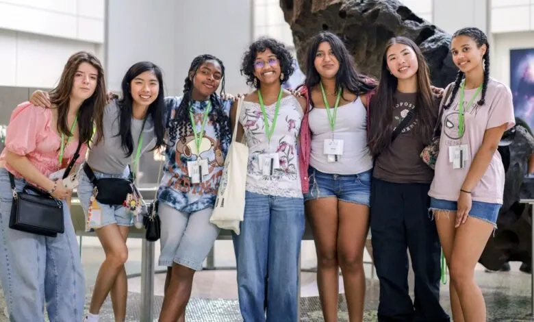 A diverse group of six teenage girls smiling and posing together in front of museum exhibits.