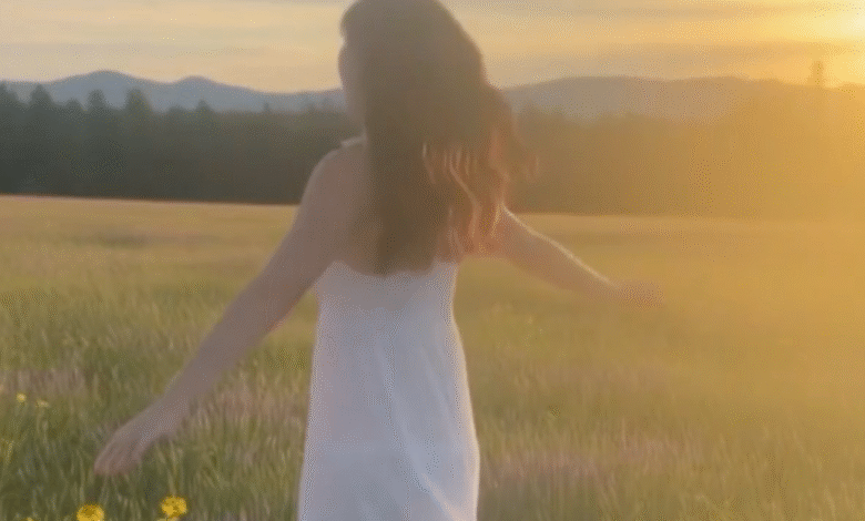Woman in white dress twirling in a sun-drenched field with mountains in the background.