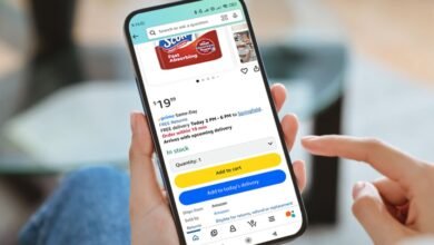Person's hand holding a smartphone displaying the Amazon app with Scott paper towels in the cart.