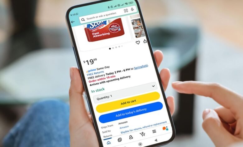 Person's hand holding a smartphone displaying the Amazon app with Scott paper towels in the cart.