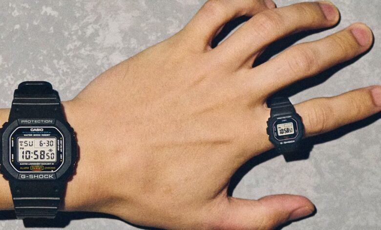 A person's hand wearing a Casio G-Shock watch on the wrist and a miniature G-Shock watch as a ring.