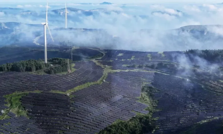 Aerial view of a vast solar farm and wind turbines on a misty mountainside.