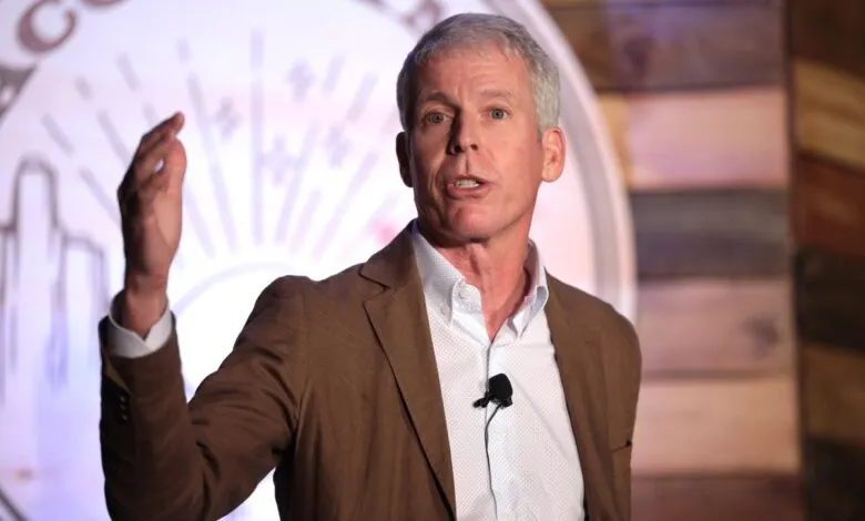Man in a brown suit jacket speaking at a conference with his hand raised.