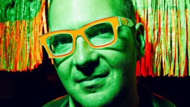 Close-up of a man wearing bright orange glasses against a backdrop of green and red fringe.