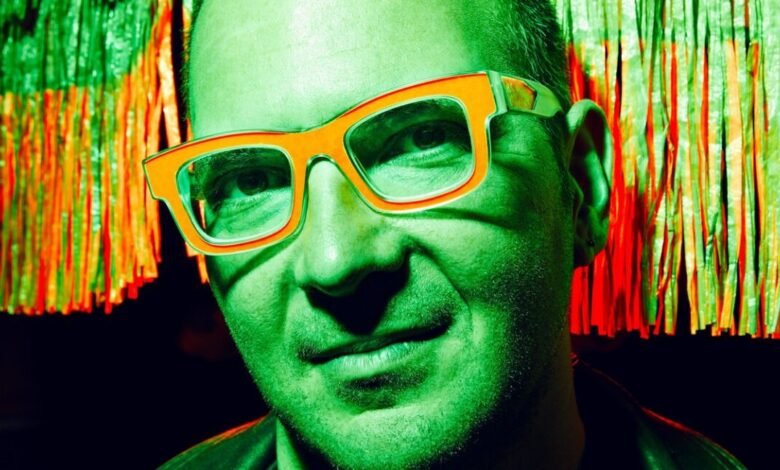 Close-up of a man wearing bright orange glasses against a backdrop of green and red fringe.