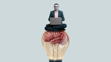 Man in suit meditating on a giant brain inside a lightbulb, working on a laptop.
