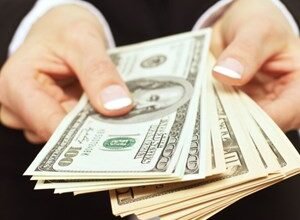 Hands holding a fan of US dollar bills, including $100 and $10 denominations.