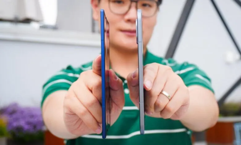 Person holding two smartphones side-by-side, one blue and one silver, for comparison.