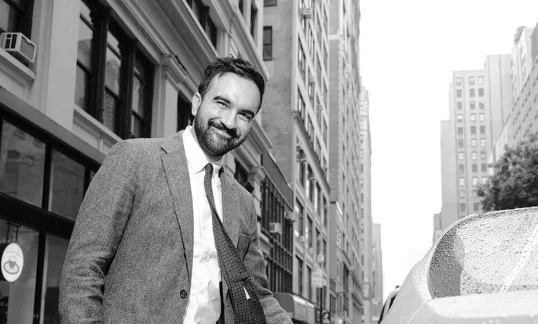 Man in suit smiling on a rainy city street with buildings and a car.