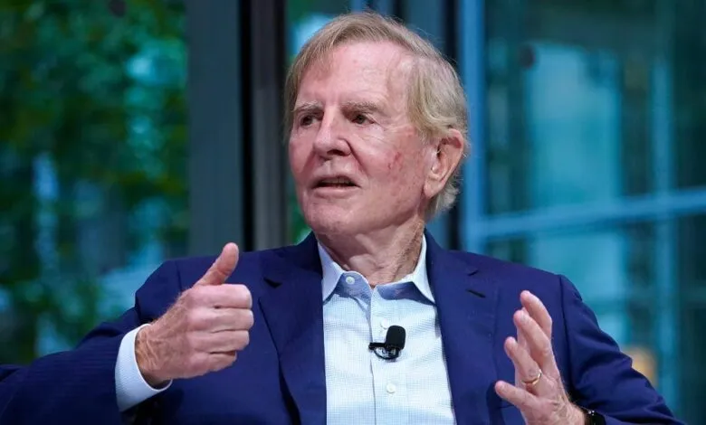 Elderly man in a suit speaking, giving a thumbs up gesture.