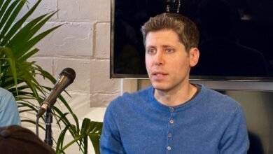 Man in blue shirt speaking into a microphone with plants and a screen in the background.
