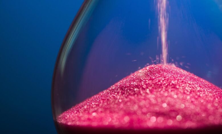 Close-up of pink glitter sand falling through an hourglass against a blue background.