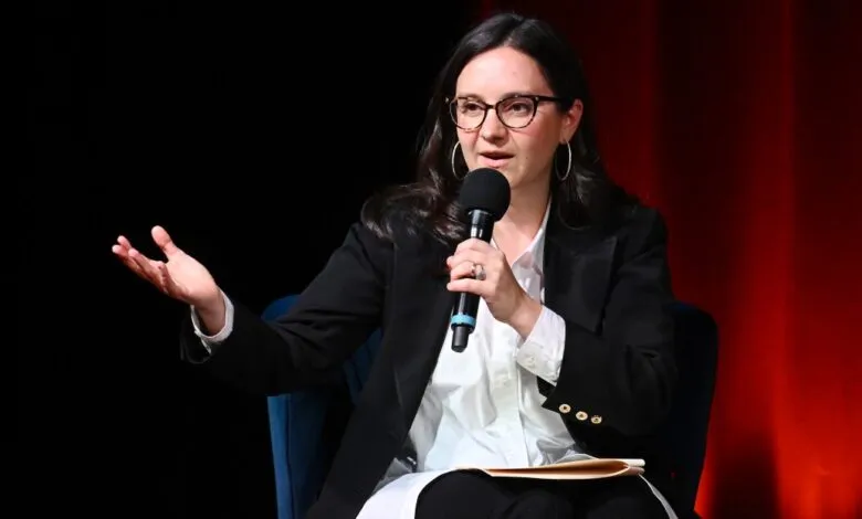 Woman in glasses speaking into a microphone, gesturing with her left hand.