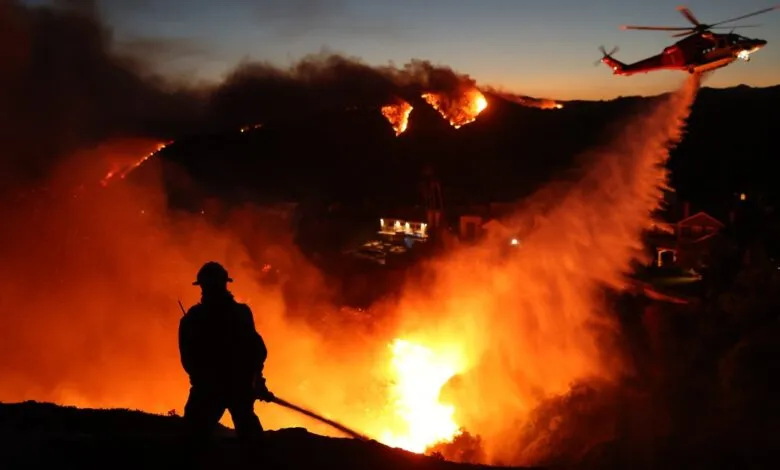 Firefighter battling a wildfire with a hose as a helicopter drops water.
