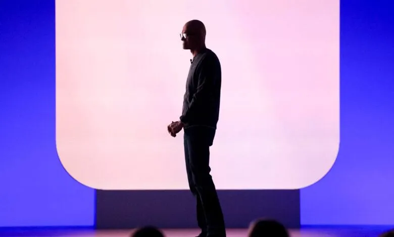 Silhouette of a bald man in glasses and casual attire on stage with a large screen.