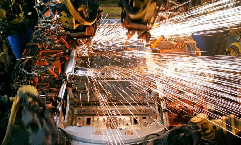 Robotic arms welding a car chassis, creating a shower of bright orange sparks.