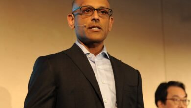 Man in a suit and glasses speaking at a conference with a microphone clipped to his collar.