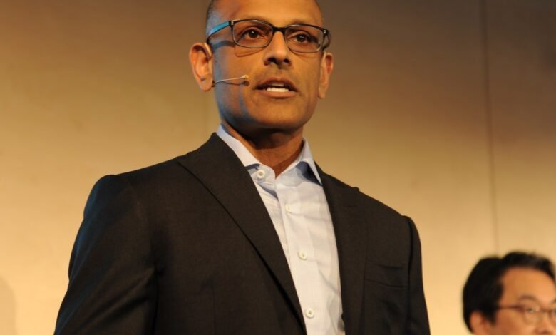 Man in a suit and glasses speaking at a conference with a microphone clipped to his collar.