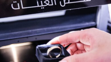 Hand holding a gold ring in a velvet box near a testing machine with Arabic text.