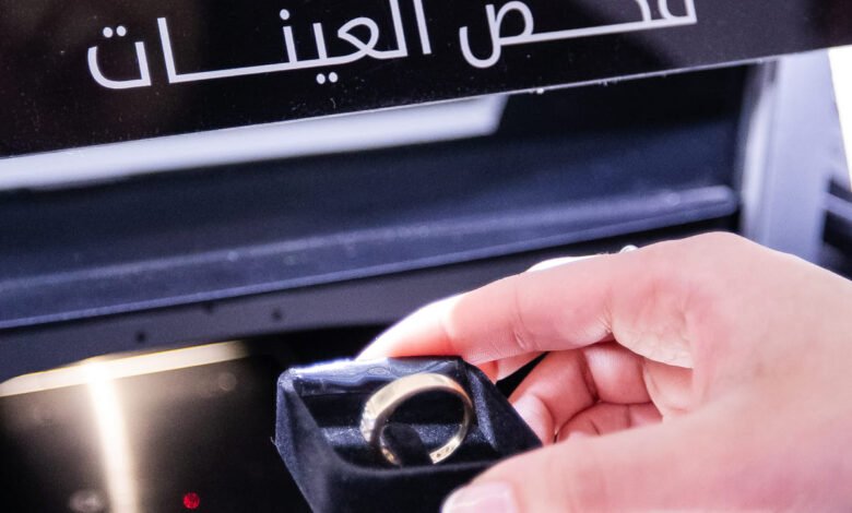 Hand holding a gold ring in a velvet box near a testing machine with Arabic text.