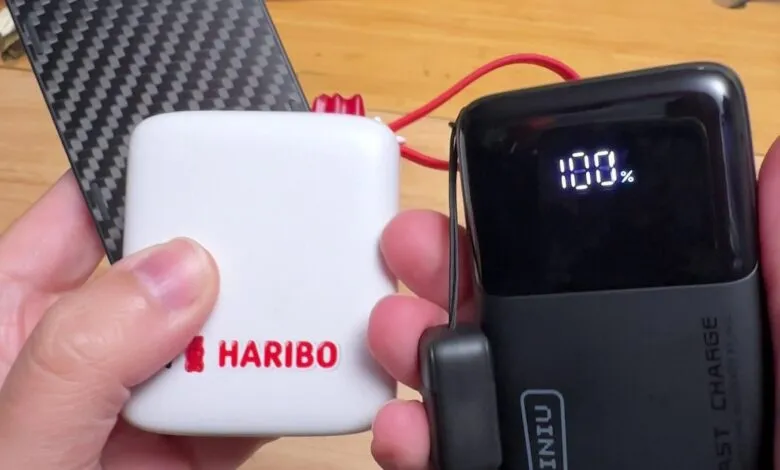 Person holding a white Haribo power bank and a black Nitecore power bank showing 100% charge.