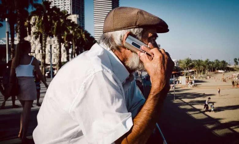 Elderly man with a cap talking on a vintage cell phone with a beach in the background.