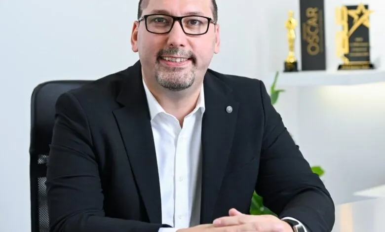 Man in a black suit and glasses smiling, seated at a desk with Oscar awards in the background.