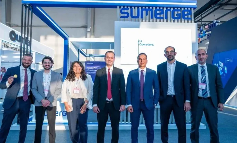 A diverse group of seven professionals posing for a photo at a business expo, with company branding in the background.