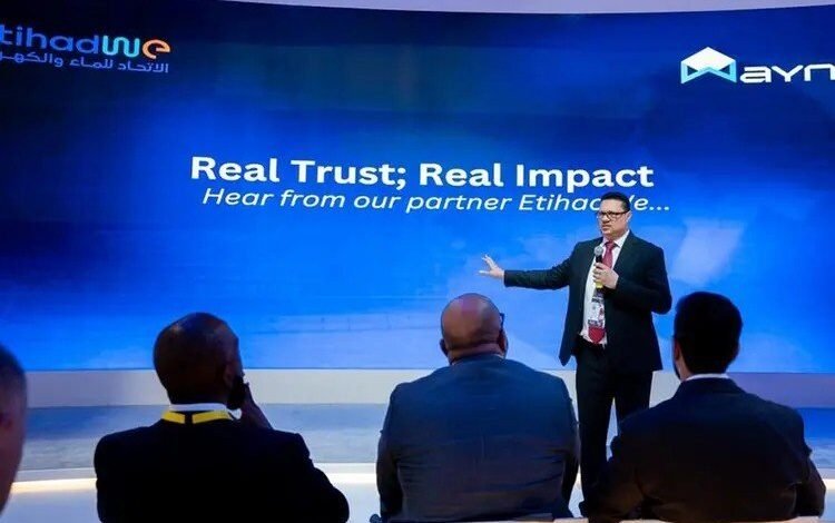 A man in a suit presents on a stage with a blue screen behind him.