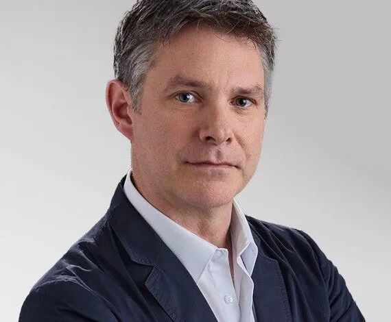 Portrait of a middle-aged man with short graying hair and blue eyes, wearing a white shirt and navy blazer.