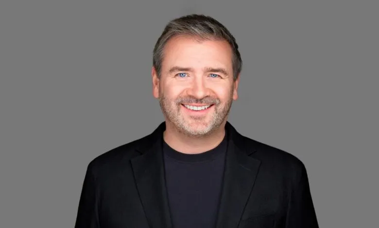 Portrait of a smiling man with short gray hair and blue eyes, wearing a black blazer.