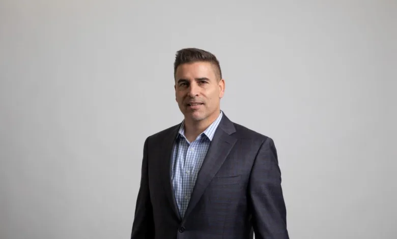 A man in a suit and checkered shirt smiles at the camera against a plain background.