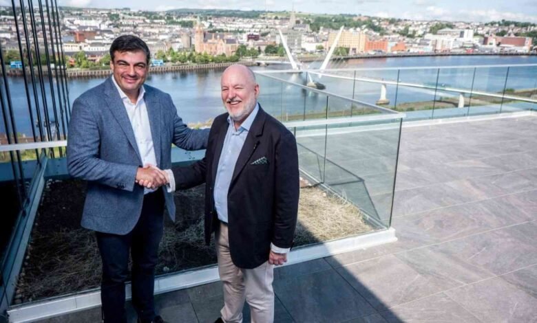 Two men in suits shaking hands on a rooftop with a city skyline and bridge in the background.