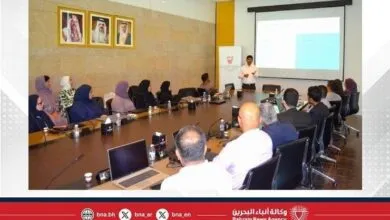A presenter stands before a projection screen, addressing an audience seated around a conference table.