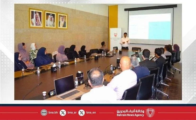 A presenter stands before a projection screen, addressing an audience seated around a conference table.
