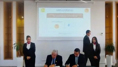 Two men in suits sign documents at a table, with two formally dressed women standing behind them.
