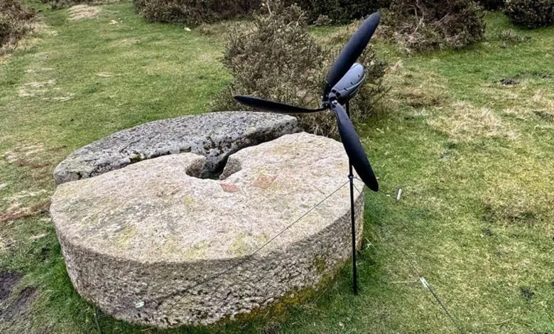 A small black wind turbine stands next to a large, weathered millstone in a grassy field.